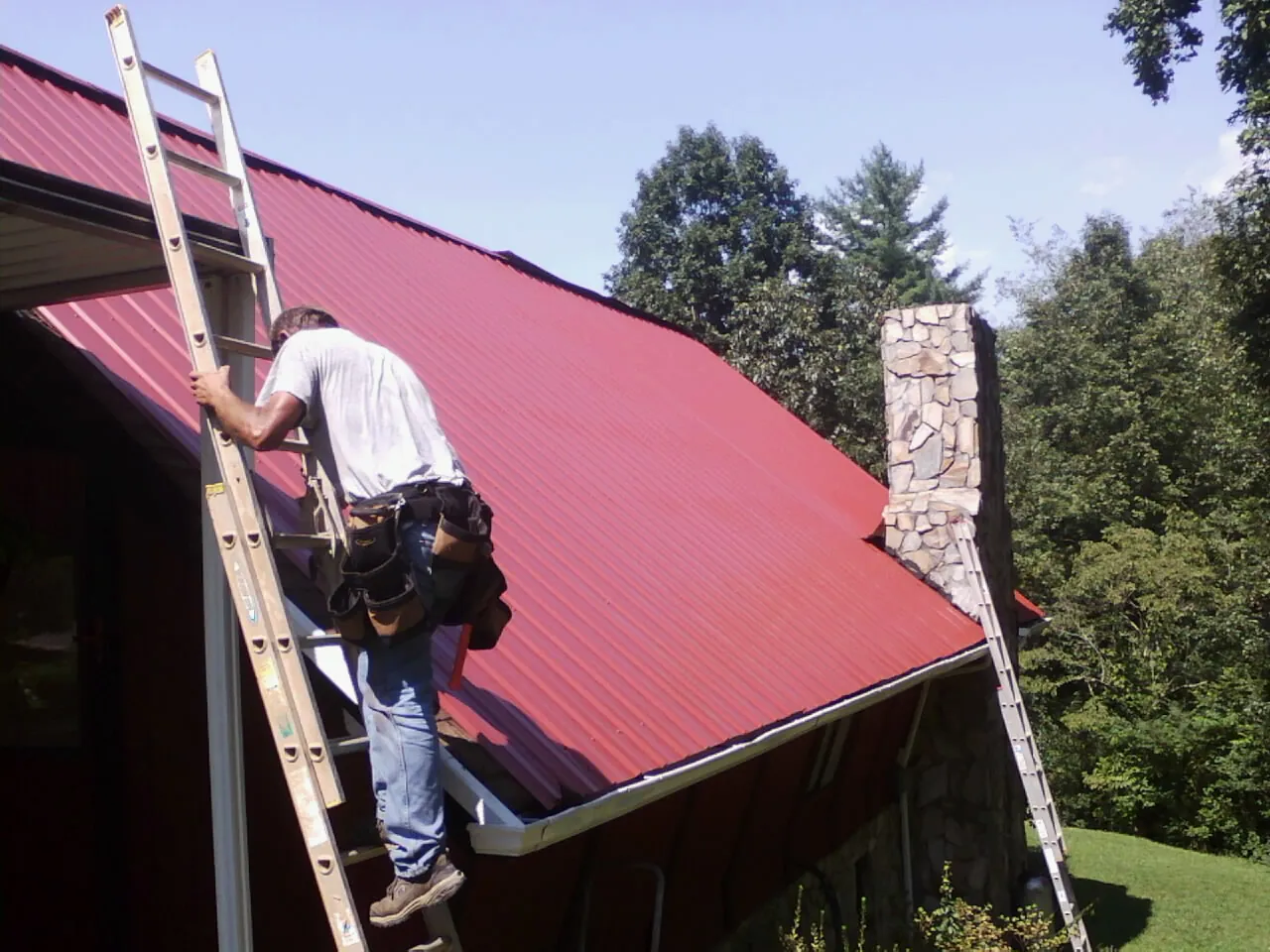 Roofer on a ladder beside a red metal roof and stone chimney — Soffit & Fascia Repair by Roofing Brothers in Wrenshall